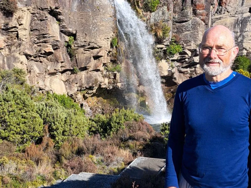 Person standing in front of a scenic waterfall with rocky cliffs and lush greenery outdoors.