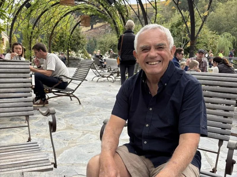 Older man with grey hair wearing a dark short-sleeved shirt and khaki shorts sitting on an outdoor wooden bench in a leafy garden café with people relaxing and socializing in the background