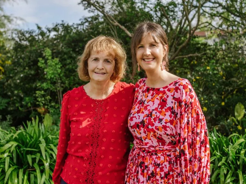 Two women standing outdoors in a garden, one wearing a solid red blouse, and the other wearing a colorful floral patterned red and pink dress, with lush greenery and trees in the background.
