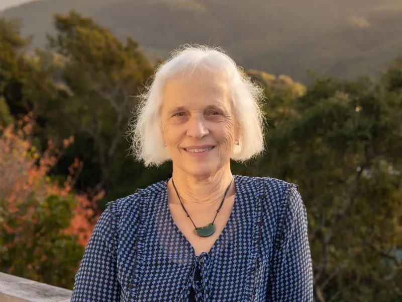 Senior woman with white hair standing outdoors in front of scenic greenery and mountains, wearing a patterned blue blouse and green stone necklace.