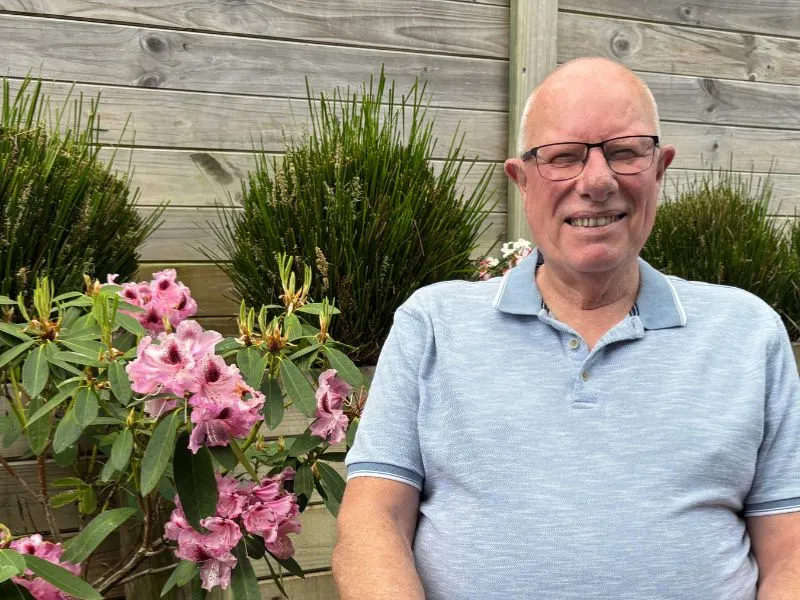 Smiling older man wearing glasses and a light blue polo shirt sitting outdoors next to blooming pink flowers and green plants with a wooden fence background.