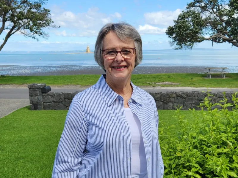 Senior woman in blue striped shirt standing in front of green grass and scenic coastal view with trees, stone wall, and blue sky.