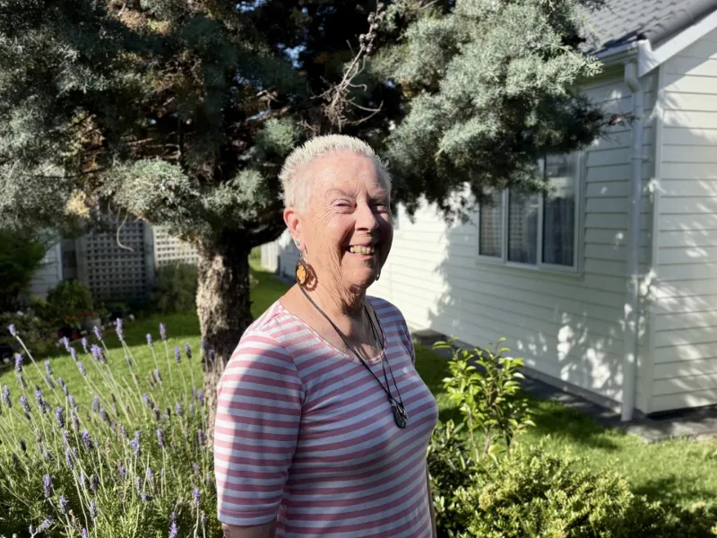 Woman with short blonde hair wearing striped shirt and necklace standing in a garden with lavender and greenery in front of a white house.