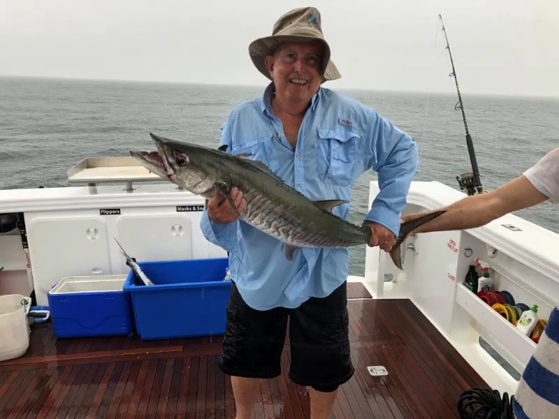 Man in blue fishing shirt and hat holding large king mackerel fish on boat with ocean background, fishing trip catch of the day.