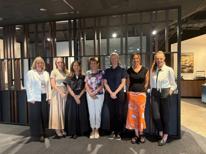 Group of colleagues standing together in a modern office lobby, team photo with diverse women in business casual clothing.