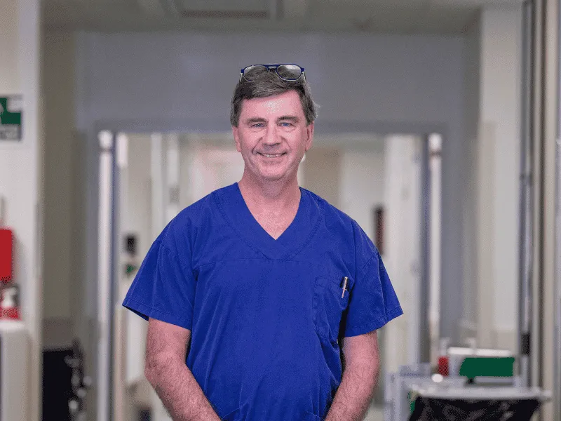 Smiling cardiologist Dr Chris Nunn in blue scrubs standing in a hospital corridor, New Zealand heart specialist supported by Heart Foundation overseas training.