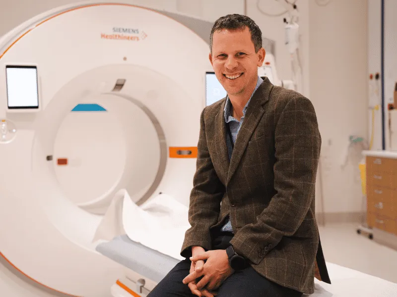 Christchurch cardiologist Dr Phil Adamson sitting in front of an advanced CT scanner, leading Heart Foundation‑supported cardiac imaging and heart disease diagnosis research in New Zealand..