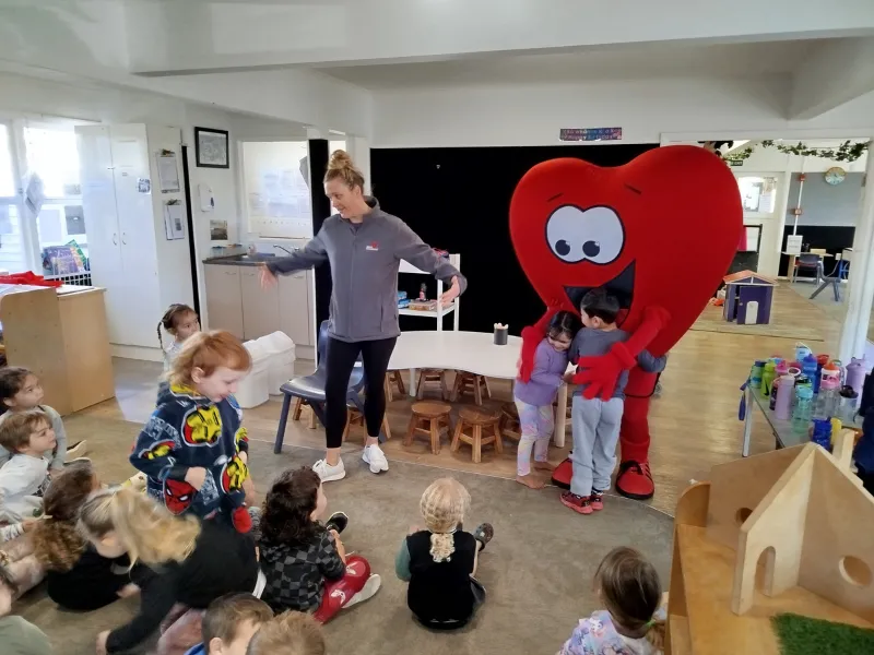 A person in a large red heart mascot costume hugs two children while another adult leads a group of young children in movement activities inside a classroom.