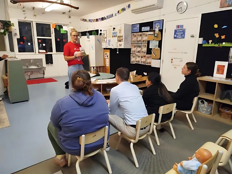 An adult stands at the front of a classroom speaking to a small group of seated adults, with posters and educational materials on the walls around them.