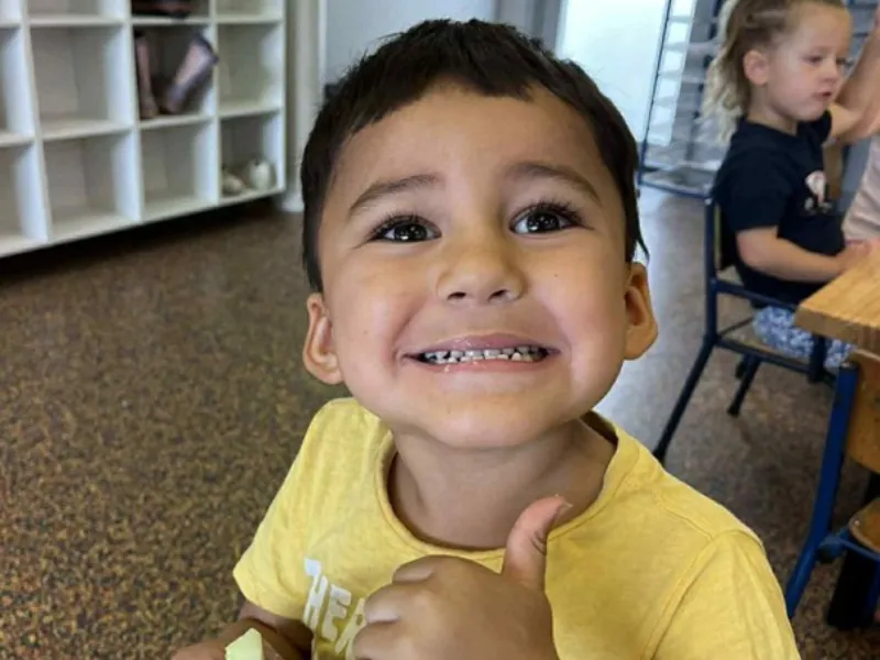 Child sitting at a classroom table giving a thumbs up while holding food, with other children and shelves visible in the background.