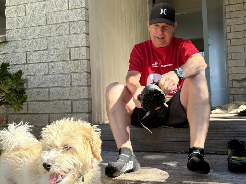 Man sitting on wooden steps in running gear tying his shoe, wearing a red Heart Foundation T‑shirt and sports watch, with a small fluffy dog in the foreground outside a brick house.