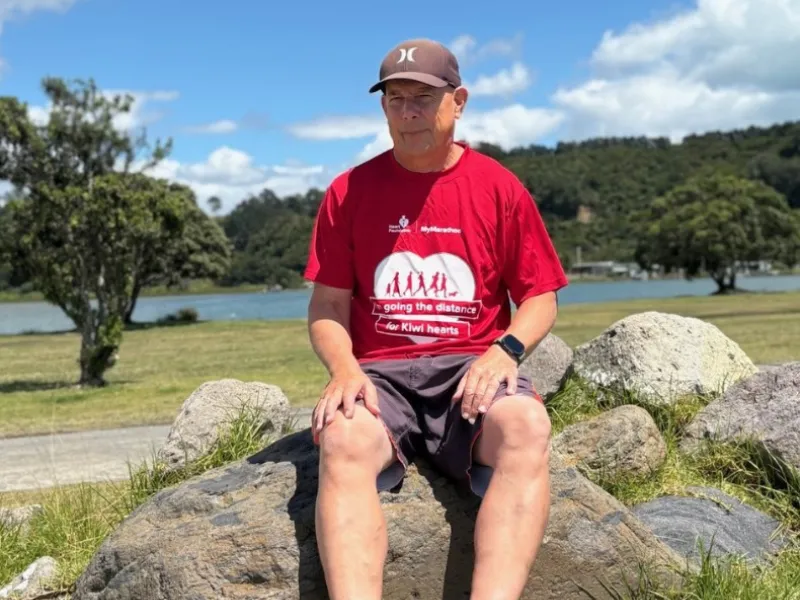Man sitting on large rocks in a park beside a lake, wearing a red Heart Foundation running T‑shirt, shorts, and a sports watch, with green hills and trees in the background.