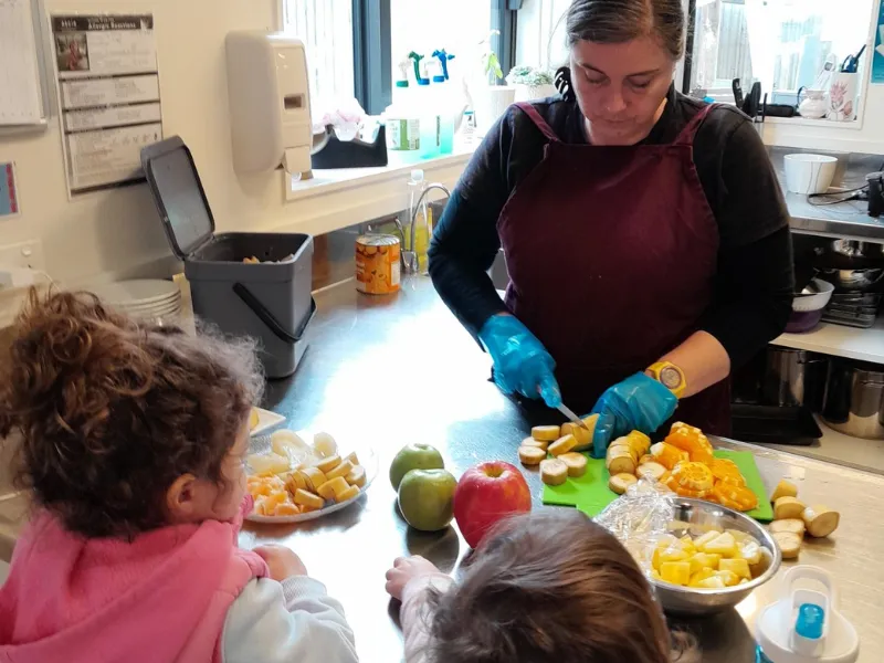 A woman wearing an apron and blue gloves slices fresh fruit on a kitchen counter while two young children watch, with plates and bowls of cut apples, bananas and other fruit in front of them.