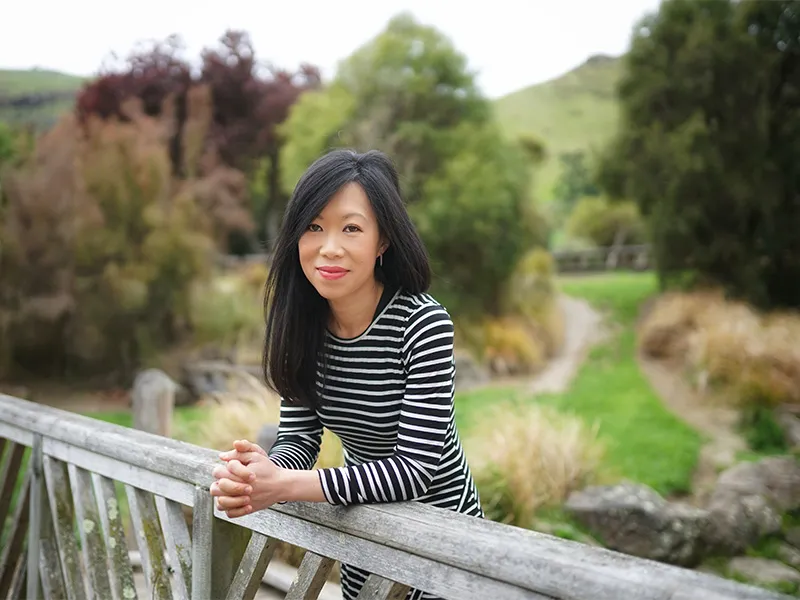 Janice Chew-Harris is a young Asian woman. She is pictured here in a rural outdoor setting. She is leaning against a wooden fence. She is wearing a black and white striped dress and bright pink lipstick.