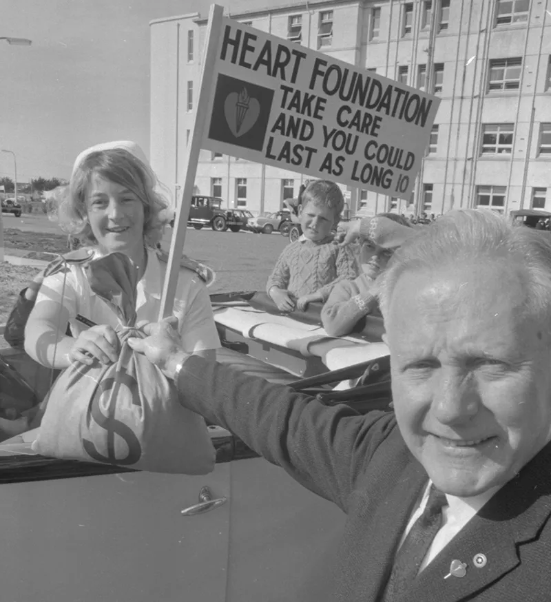 Black and white photo of Jocelyn Bell sitting in a vintage fire truck holding a Heart Foundation banner. She is handing a bag of money over to an important looking official. 