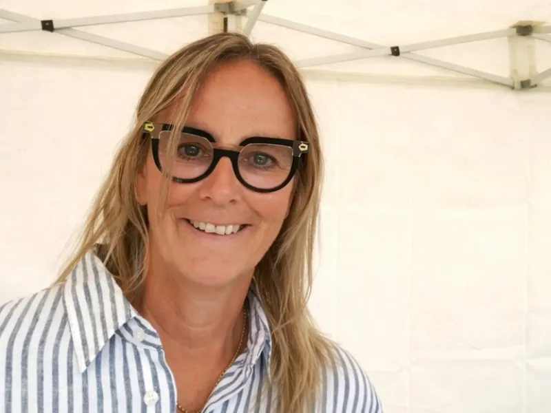 Middle‑aged woman with long blonde hair wearing a blue and white striped shirt standing under a white canopy tent.