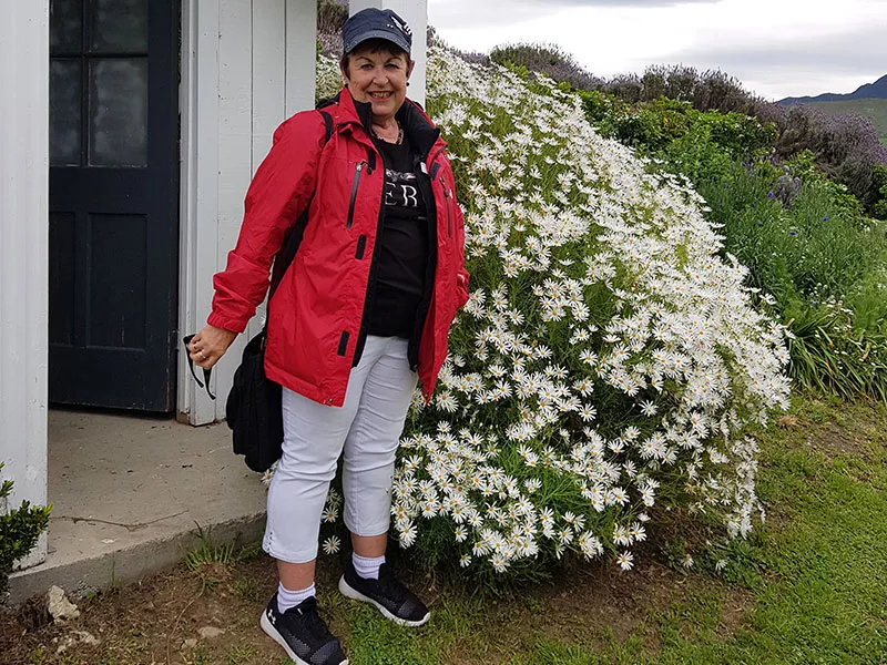 Maggie stands in the porchway of an early settler building, with flowering shrubbery to the side. She wears white trousers and a red waterproof jacket.