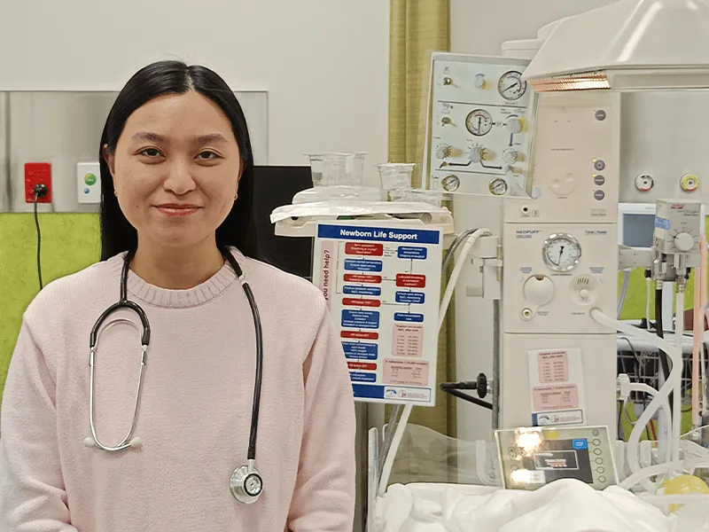 Phuong Bui - with a stethoscope standing in a medical room next to newborn life support equipment.