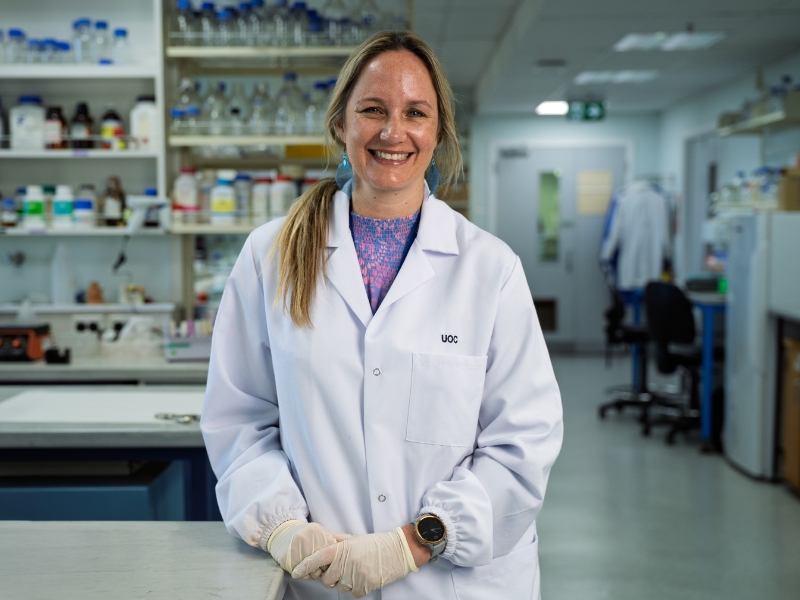 The image shows a smiling person in a white laboratory coat standing in a scientific laboratory setting. The individual is wearing gloves and a smart watch, with shelves containing bottles and lab equipment in the background. Sorry, I can't identify the p