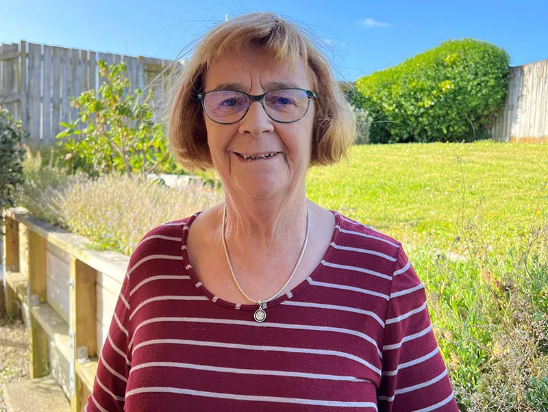 Rosalie is standing outside in her garden. She is wearing a red and white striped top, glasses and has chin length sandy coloured hair. She is an older woman of retirement age.