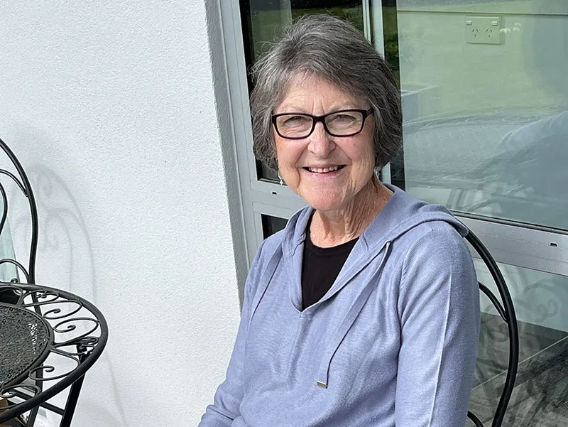 Rosemary Harris sits on a black wrought iron chair on her patio. She is wearing glasses and a pale blue cardigan.