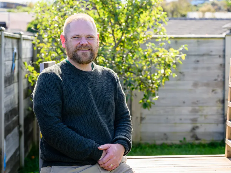 Man sitting on wooden deck in sunny backyard garden with green tree and fence in background.