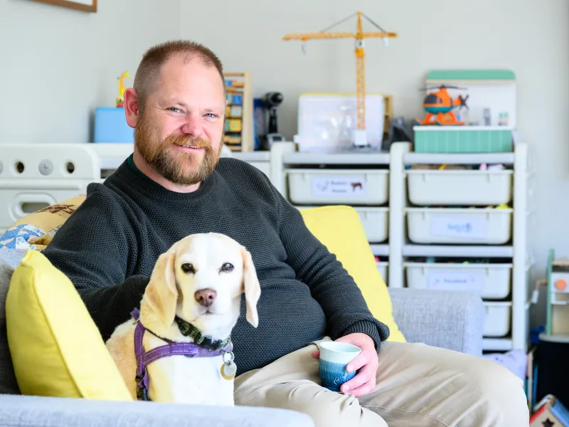 Man relaxing on couch at home with beagle dog, holding a coffee cup in a cosy living room.