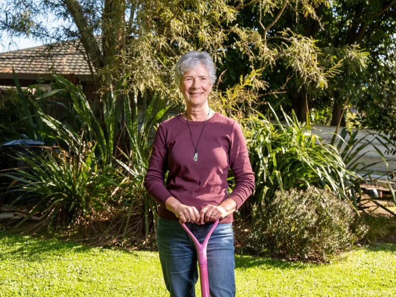 Older woman standing in a sunny backyard garden holding a pink rake, staying active outdoors for heart health and healthy ageing.