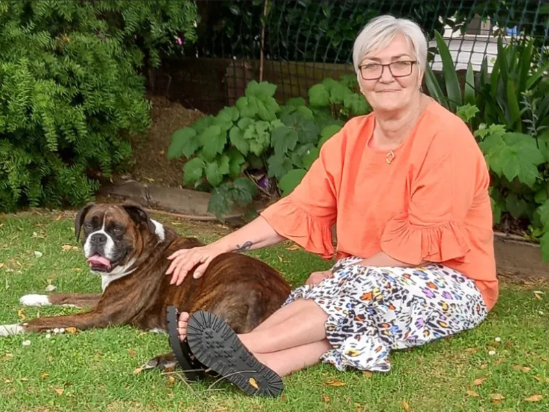 Older woman sitting on the grass in a garden with a large brown and white boxer dog lying beside her, resting her hand on the dog’s back.