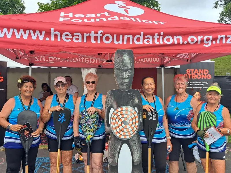 A group of six women in blue athletic outfits holding paddles stand smiling in front of a Heart Foundation tent, posing for the Stand Strong campaign