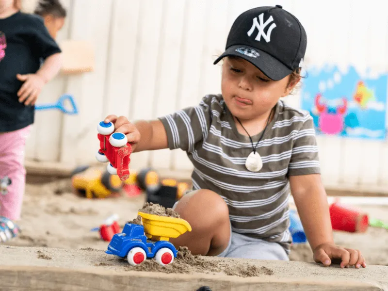 A young child wearing a striped shirt and black cap plays intently in a sandpit, using a toy excavator to load sand into a colourful toy dump truck. The child appears focused and playful, with their tongue slightly out in concentration. Other toys and chi