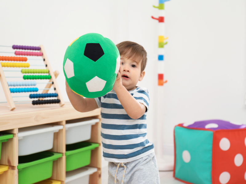 Young child playing with a green plush soccer ball in a colorful playroom with educational toys and storage bins.
