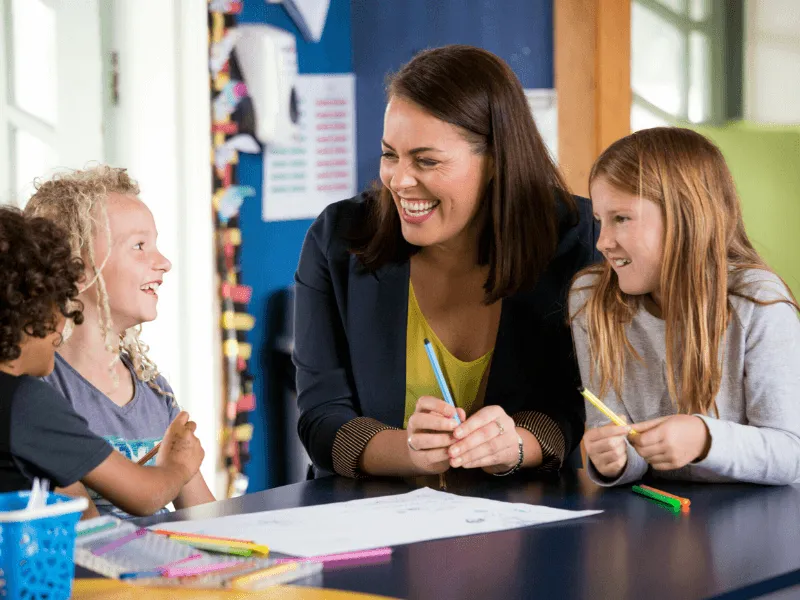 A smiling woman sits at a classroom table, engaging warmly with three children—two girls and one boy—while they draw with colorful markers. The group appears cheerful and connected, suggesting a nurturing and inclusive learning environment.