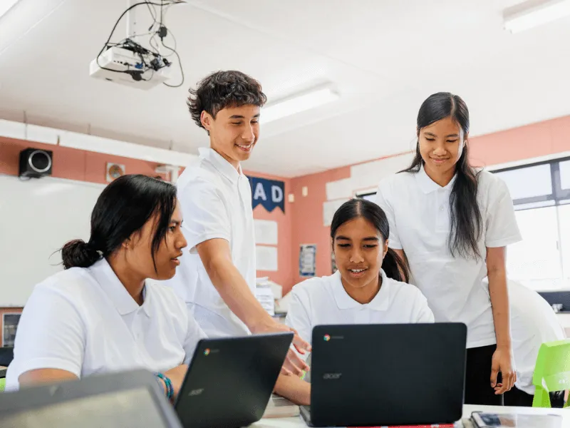 Four high school students in white polo shirts collaborate around laptops in a bright classroom. One student stands and points at a screen while others sit, engaged in discussion and learning.