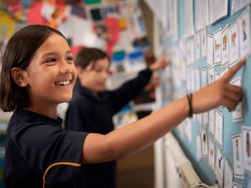 A smiling young student points at a display of cards on a classroom wall, with another student in the background doing the same. The wall is covered in colorful educational materials, creating a lively and engaging learning environment.