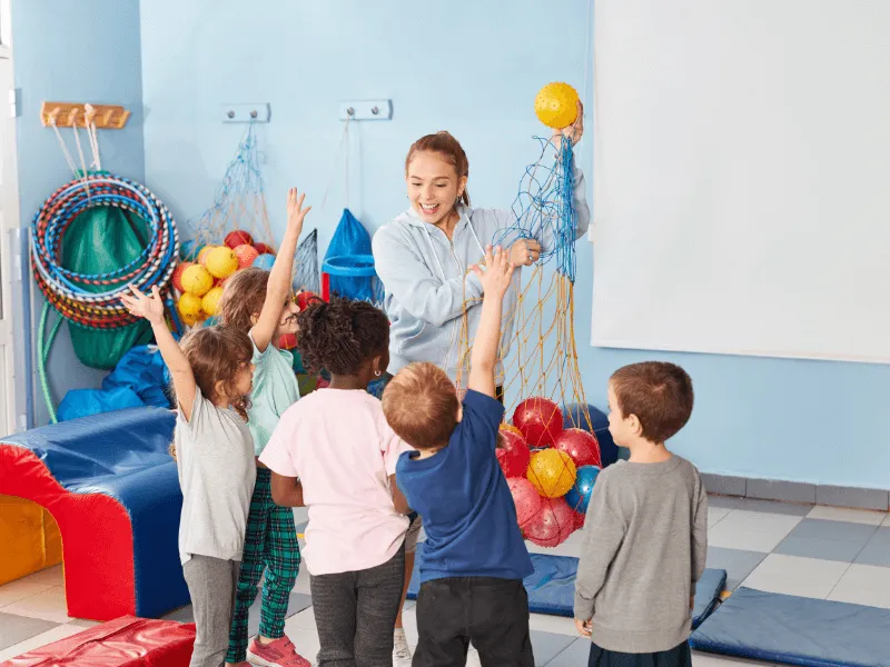 A group of young children excitedly reach up as a teacher hands out balls from a net in a colorful indoor playroom filled with soft play equipment and hoops.