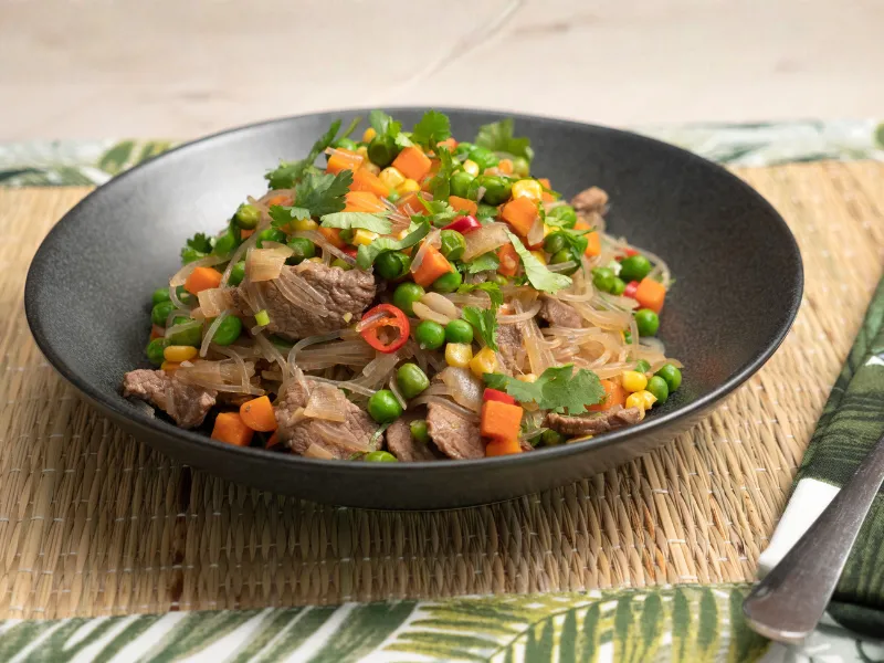 Black bowl filled with glass noodles, lean beef, mixed vegetables like carrots, peas, corn, and topped with fresh coriander, served on woven placemat.