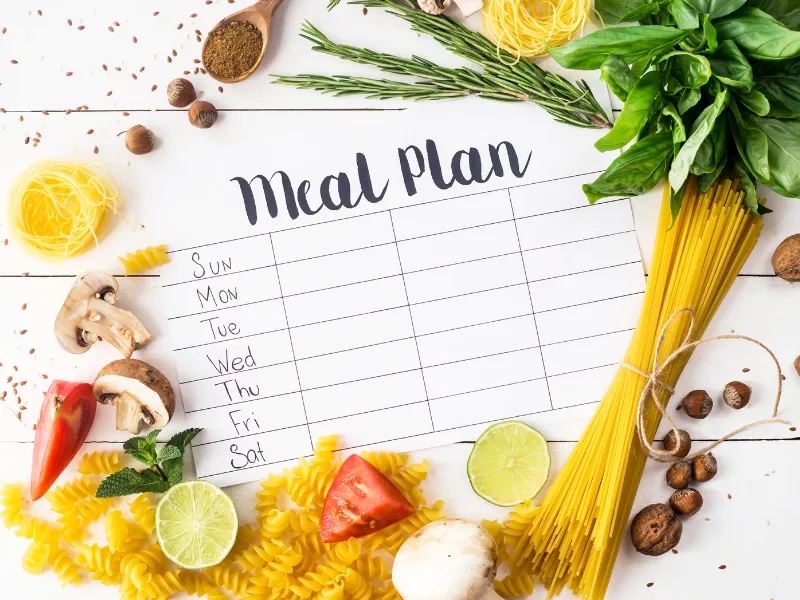 Weekly meal plan template surrounded by fresh ingredients like pasta, mushrooms, lime, tomato, leafy greens, and herbs, for healthy recipe planning.