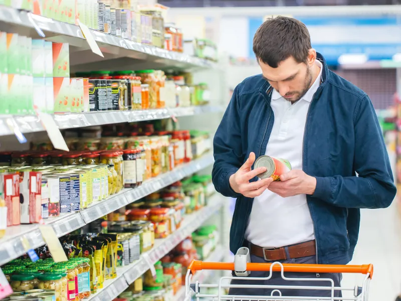Man shopping in supermarket reads label on canned food, checking ingredient or nutrition information in grocery aisle with shelves of packaged products.