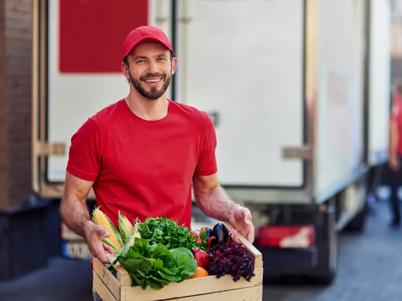 Delivery worker in red uniform holding a crate filled with fresh vegetables like corn, lettuce, tomatoes, and leafy greens in front of refrigerated truck.