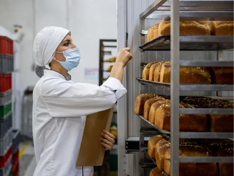 Bakery worker in uniform inspecting fresh bread loaves on racks, wearing mask and hairnet, quality control in commercial bakery production facility