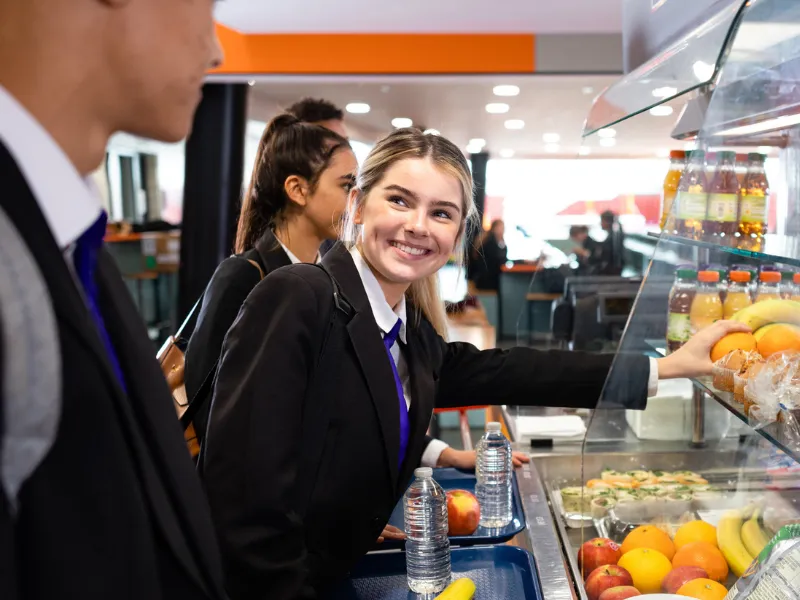 Students in school uniforms selecting fresh fruit, sandwiches, and bottled drinks from a cafeteria counter filled with healthy menu options