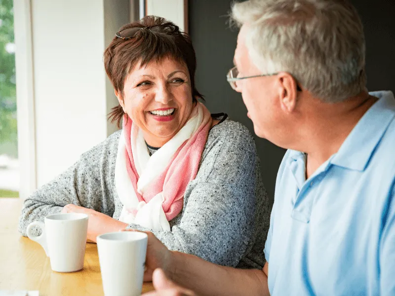 Two people sitting at a table with coffee mugs, having a conversation in a bright room.