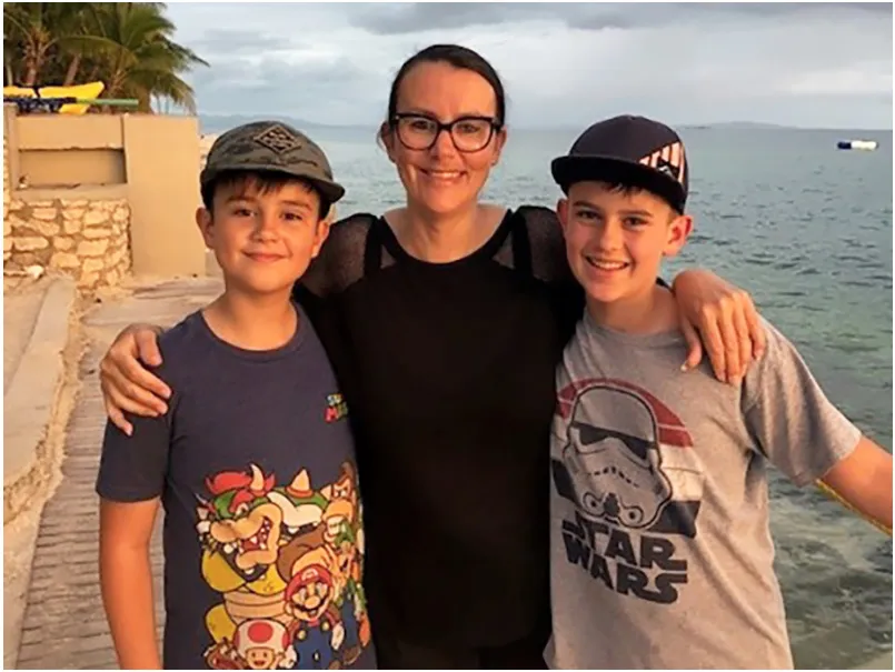 Tanya Neeley stands, smiling, with her arms around the shoulders of her two sons. The sun is setting and the sea is visible behind them.