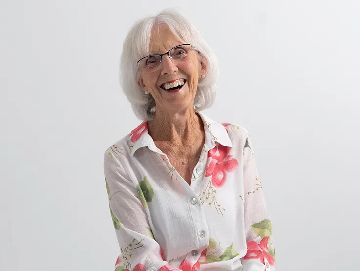 Claire is a senior lady wearing a white and pink floral shirt. She is seated on a stool and looking into the camera smiling.