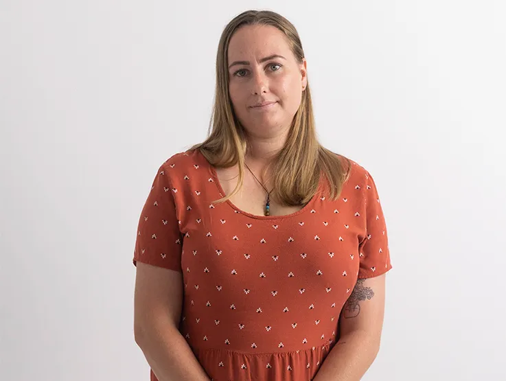 Kerri is a young woman in her twenties. She has long, fair hair and is wearing a pretty summer dress in a peach colour. She is standing in front of the camera with a ghost of a smile on her lips, like she is a bit shy to be photographed.