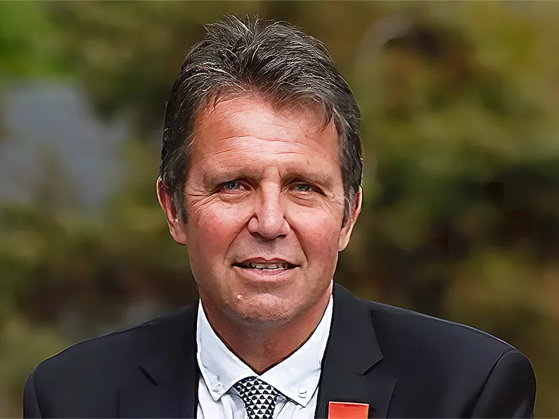 Portrait of Dr John Mayhew. He is wearing a dark blue suit, white shirt and a red pocket square. The background is burred but appears to be foliage.