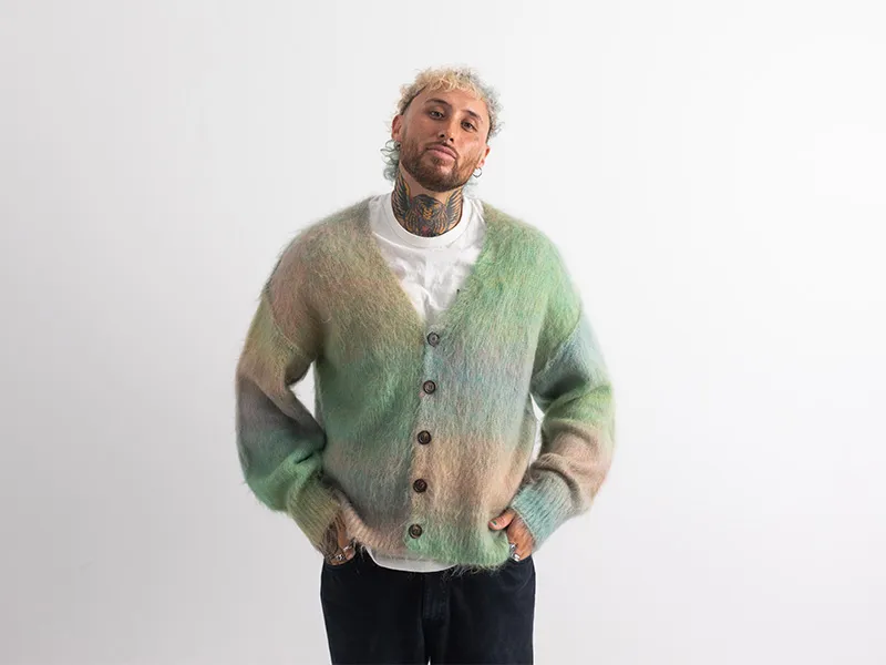 Lance Savali is wearing a pastel fluffy cardigan and is facing towards the camera looking directly forward, with his hands in his pockets. He is not smiling.