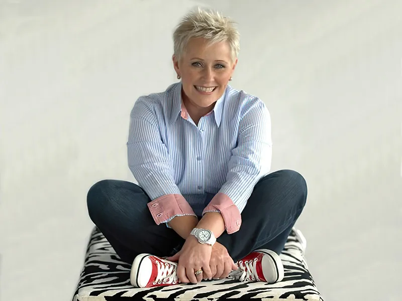 Suzanne is sitting cross-legged upon a cushion in a black and white zebra print. She is wearing blue denim jeans, with a pale blue button down shirt and bright converse sneakers on her feet. She is smiling broadly.