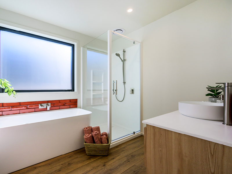 Stylish bathroom with freestanding bathtub, glass shower, wooden vanity, and red tile accent in a modern Wanaka property